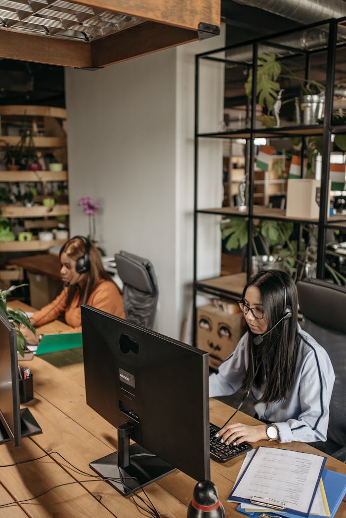 Two women in headsets working diligently in a modern office space with green plants and industrial design elements.
